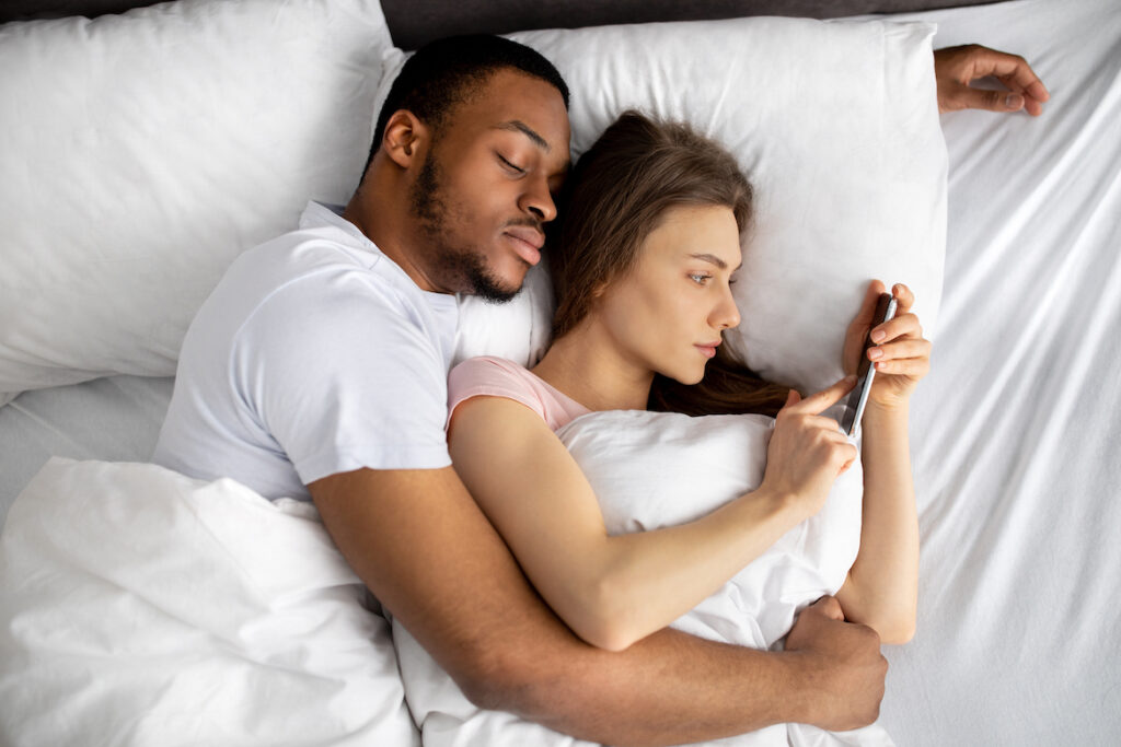 Young woman lying on bed in arms of black husband while using mobile phone